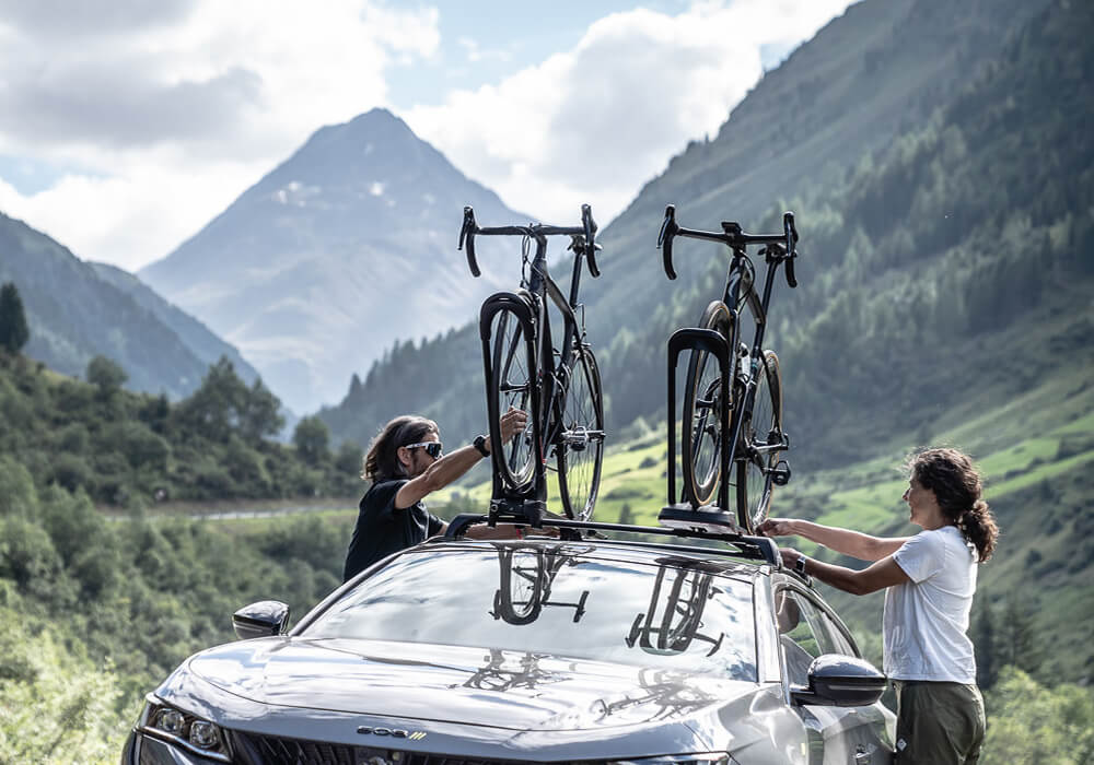 A couple secure their bikes to the roof top of their car in front of a mountain background