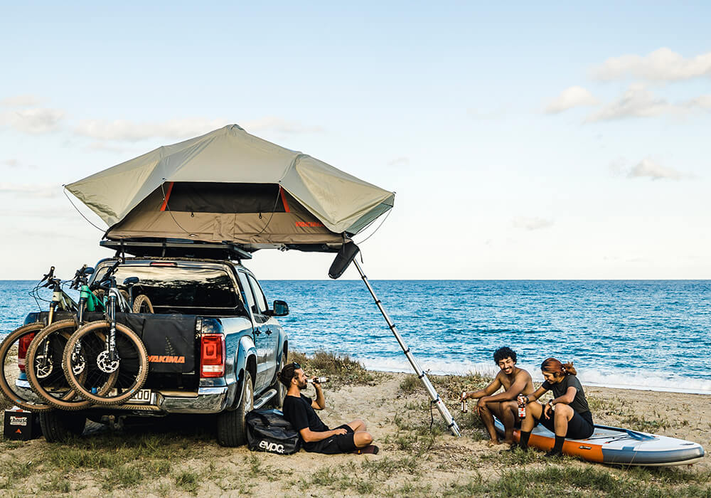 Three friends sit on a beach next to a pickup truck fitted with a roof tent with three mountain bikes in the pickup bed